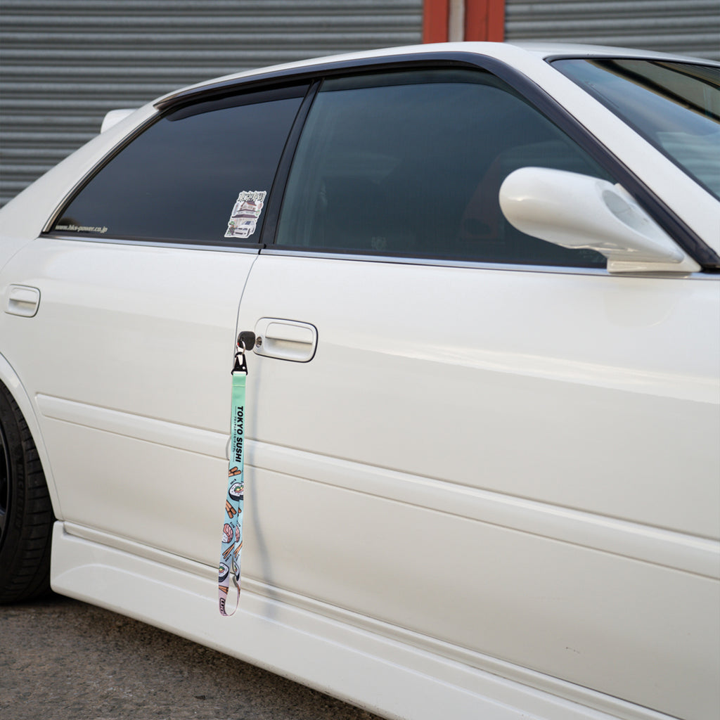 A close-up of a white car’s side door features the Blossom Automotive 'Tokyo Sushi Collection’ Lanyard hanging from the handle, with a sticker on the rear window. The car is parked beside a corrugated metal wall.