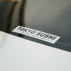 A close-up of the Blossom Automotive 'Tokyo Sushi' Slap Sticker on a car window shows its bold logo and subtle Japanese text below, with a softly blurred background.
