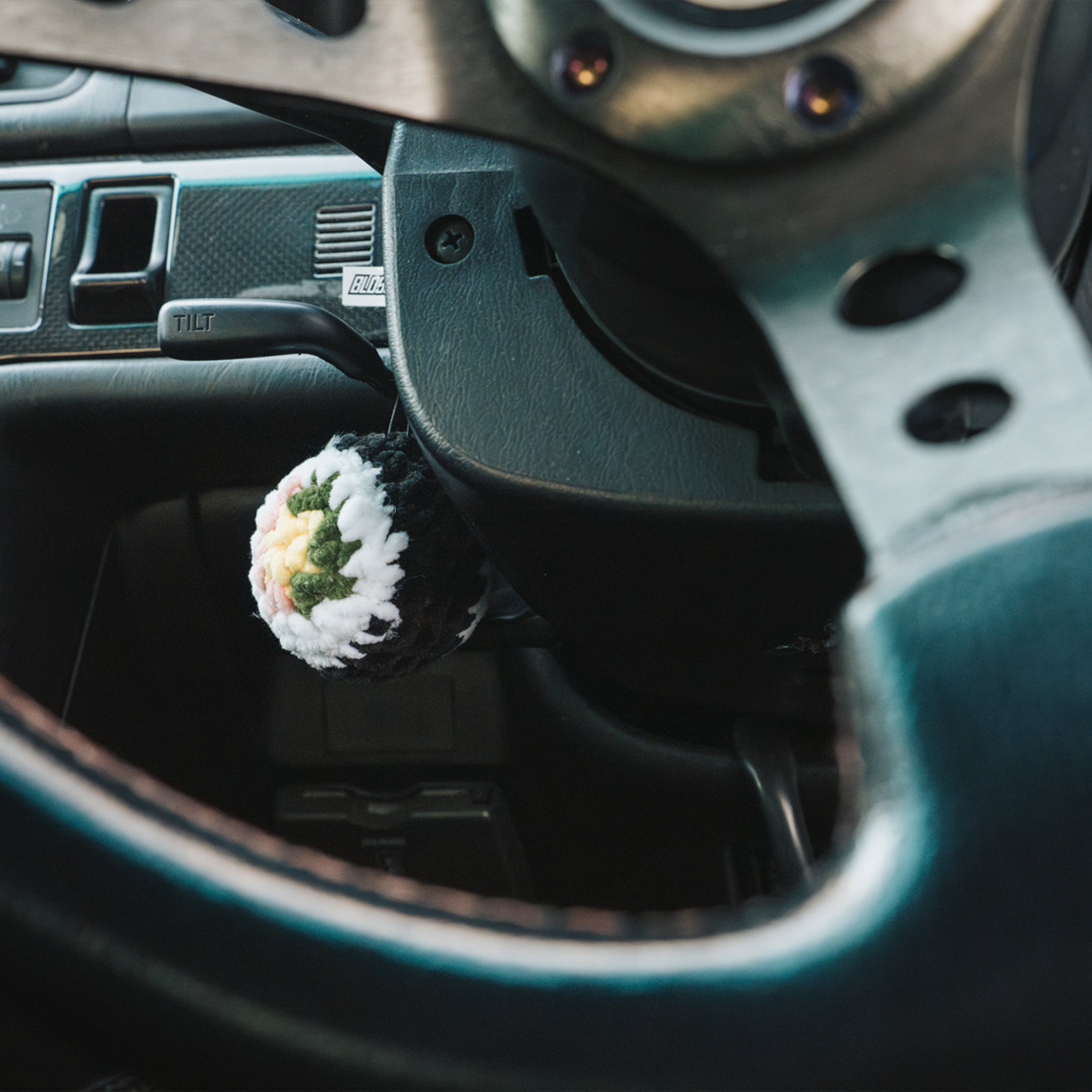 A close-up of a car steering wheel with the Blossom Automotive Squishy Sushi - Car Mirror Hanger, shaped like a sushi roll, hanging from the ignition switch. The dashboard and control switches are visible in the background.