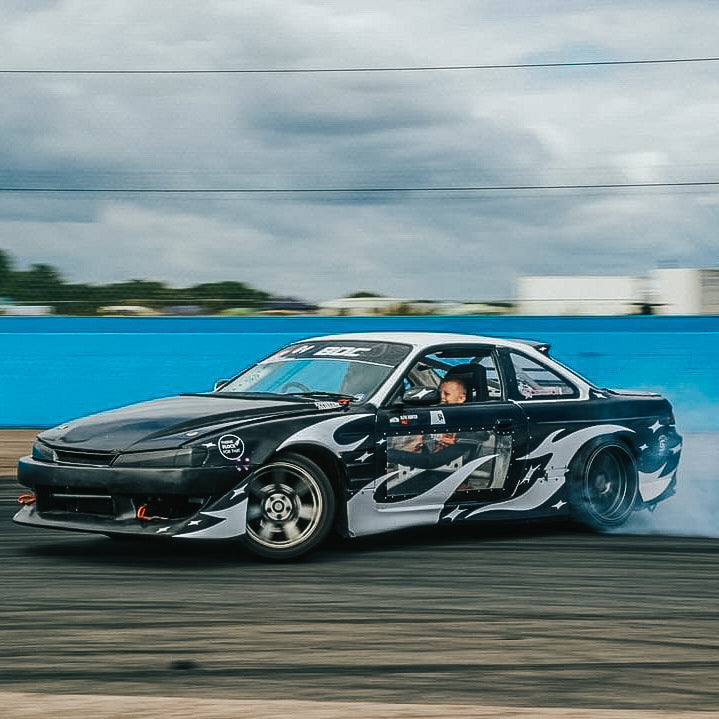A Blossom Automotive DIY 'Zoku Style' Tribal livery sports car drifts on a track, its customizable black and white decals highlighted as tire smoke rises near a blue barrier under cloudy skies.
