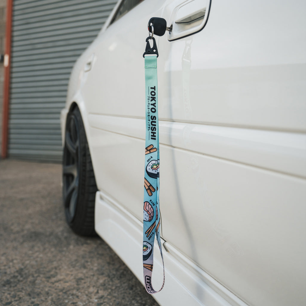A Blossom Automotive ‘Tokyo Sushi Collection’ Lanyard, featuring vibrant sushi illustrations, hangs from the door handle of a white car parked beside a corrugated metal wall.