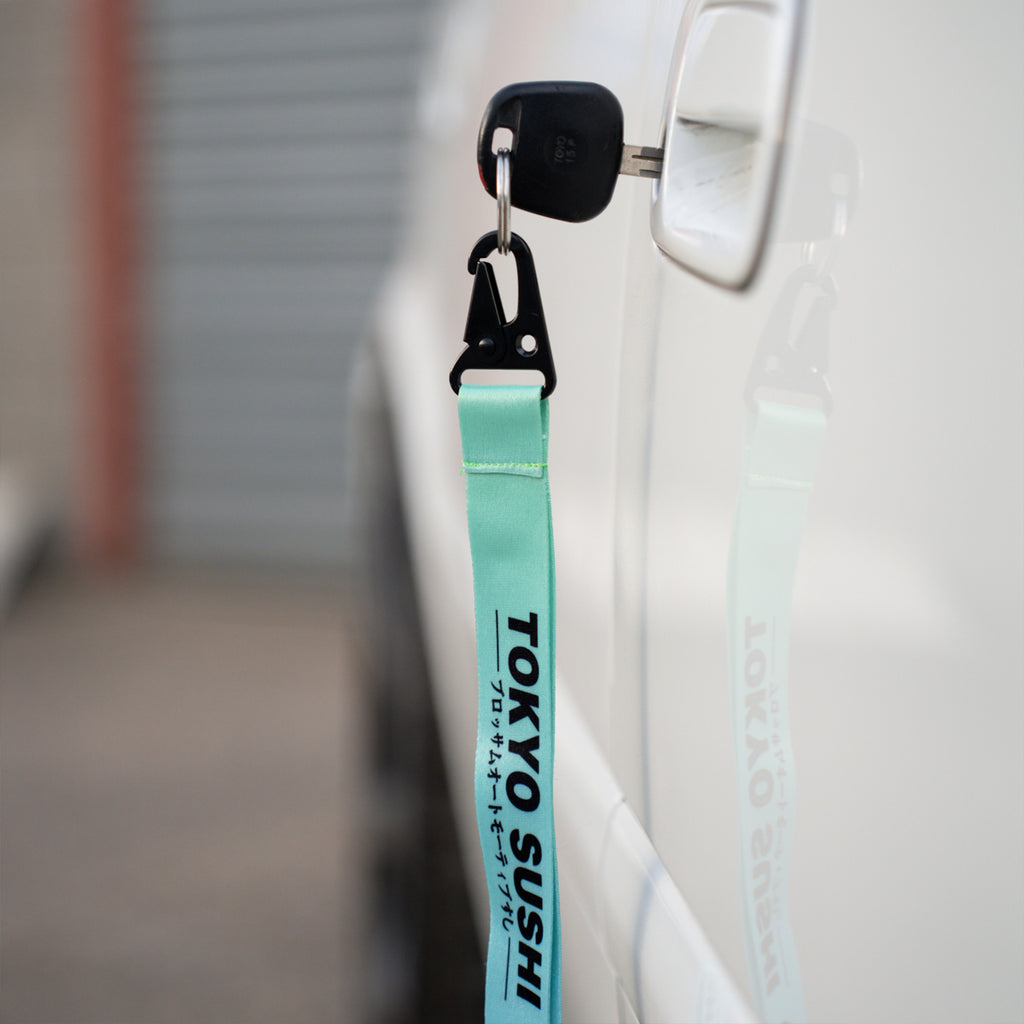A car key with a turquoise Blossom Automotive ‘Tokyo Sushi Collection’ lanyard, featuring English and Japanese text, is inserted into the door of a white vehicle, its reflection visible on the car’s surface.