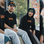 Two men sit outdoors by a metal fence, both wearing matching black ‘The Skate Collection’ Caps from Blossom Automotive. One sports a T-shirt and light jeans; the other, a hoodie and dark pants. A skateboard leans against the fence behind them.