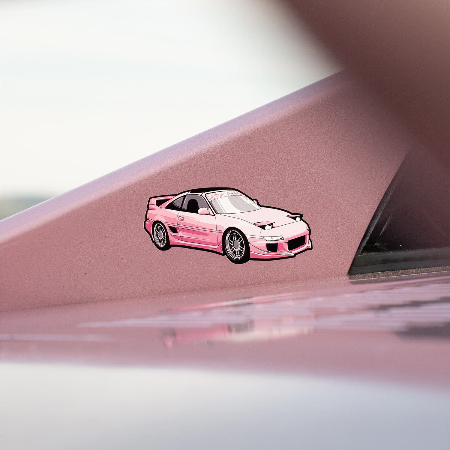 A Blossom Automotive MR2 - Slap Sticker, featuring a pink and white cartoon design, decorates the smooth pink surface of a vehicle, with part of the car’s structure blurred in the foreground.