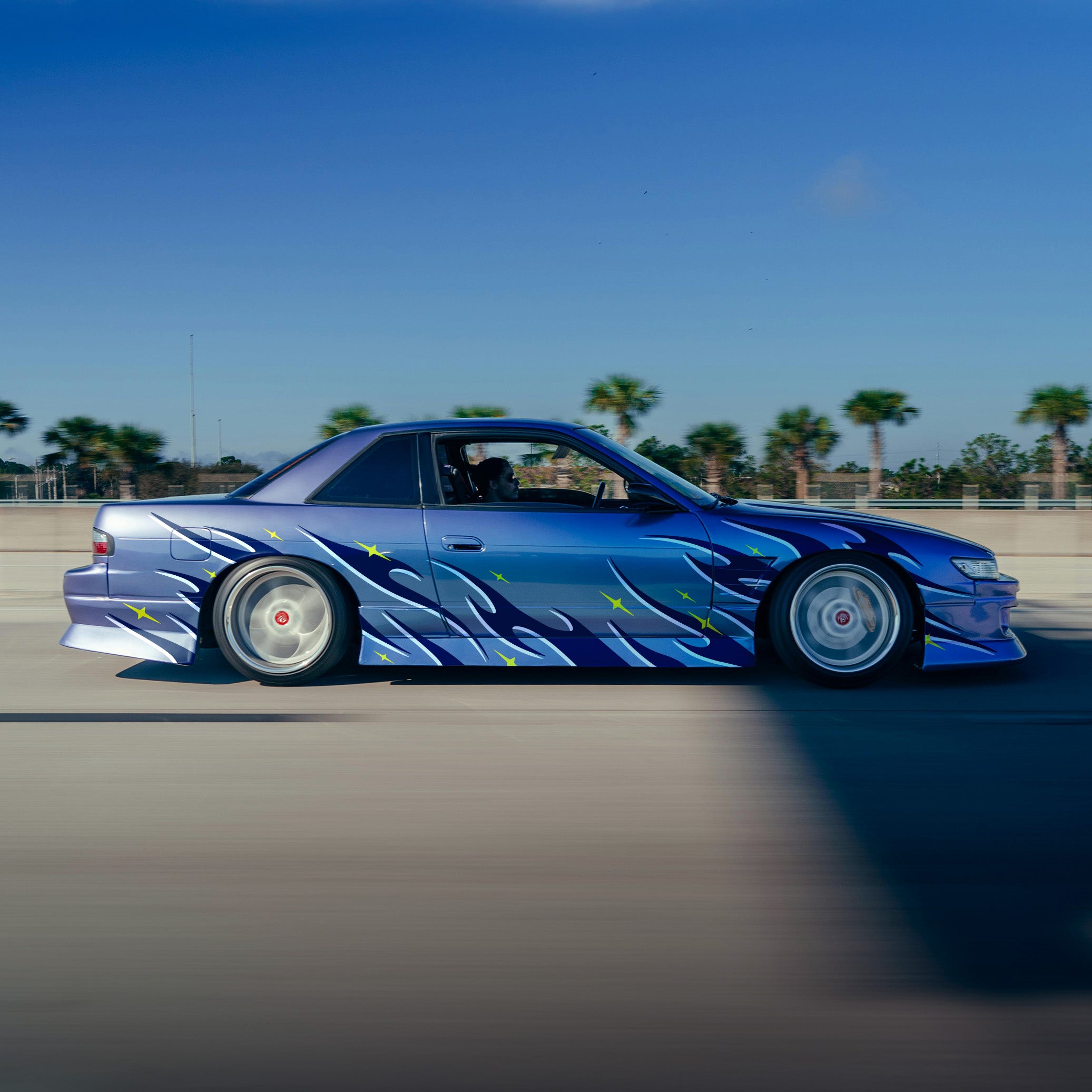 Blue car with flame design on a road with palm trees in the background