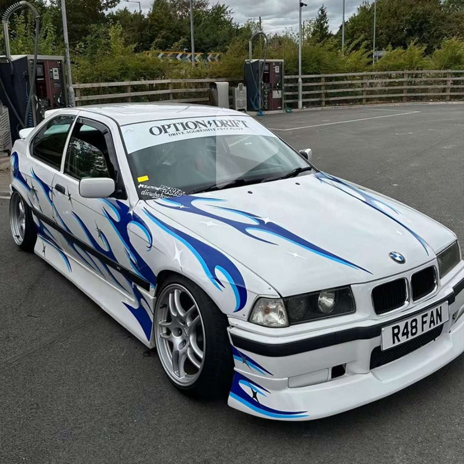 White BMW car with blue flames on a track, featuring the Blossom Enra livery.