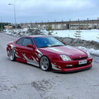 Red sports car with a Blossom Zoku livery with snow-covered ground and buildings in the background