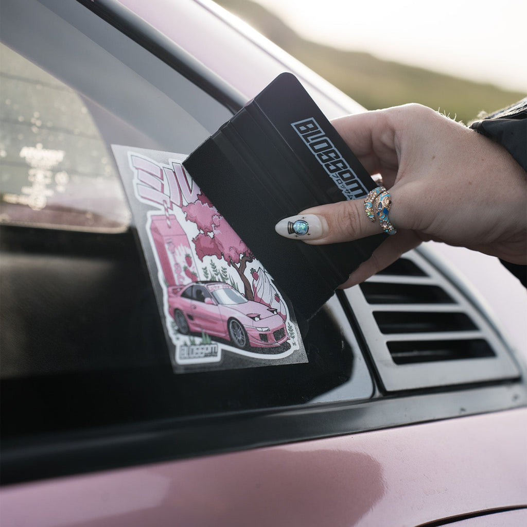 A person with painted nails and rings uses the Blossom Automotive Blossom Felt Edge Application Squeegee - Black to smooth a pink car-themed sticker onto the rear window of a pink car.