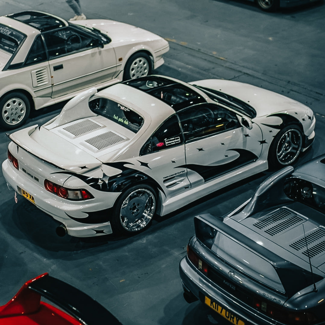A white sports car featuring Blossom Automotive’s DIY 'Zoku Style' Tribal Universal CUSTOMISABLE Livery is parked indoors among similar cars, such as a white coupe and a silver car, at what appears to be a car show or exhibition.