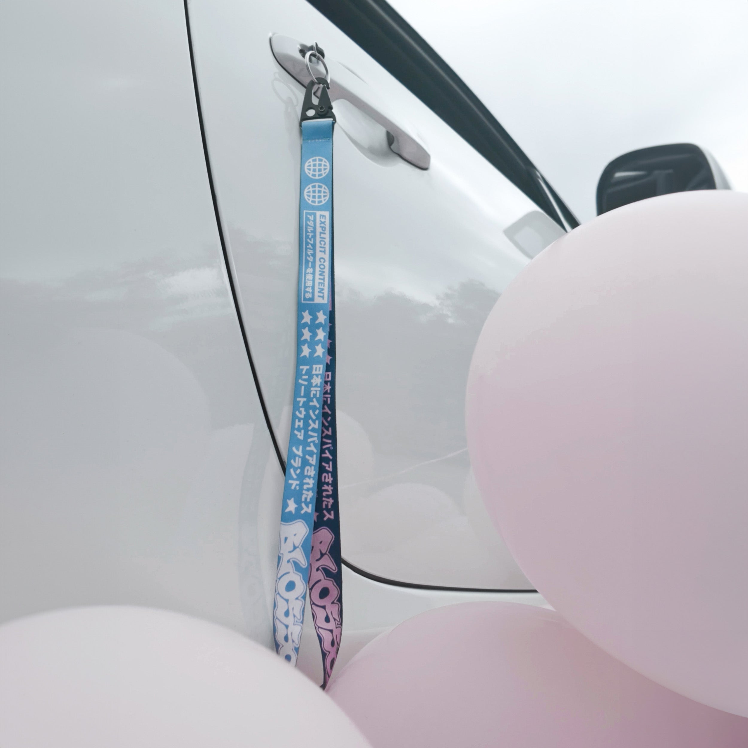 A 'The Bubblegum Collection’ Lanyard by Blossom Automotive with Japanese text in blue and purple hangs from a silver car door handle, surrounded by pink balloons.