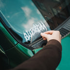 A person’s hand applies the Blossom Skate Logo - Die Cut Sticker by Blossom Automotive to the windshield of a green car.