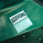 Two Blossom Skate Logo - Die Cut Stickers by Blossom Automotive, one black on white and one white on blue, are placed on the glossy green surface of a car.