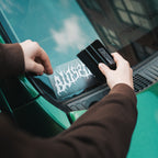 A person applies the Blossom Skate Logo - Die Cut Sticker by Blossom Automotive, featuring jagged, flame-like white lettering, to the windshield of a green car using a black plastic tool.