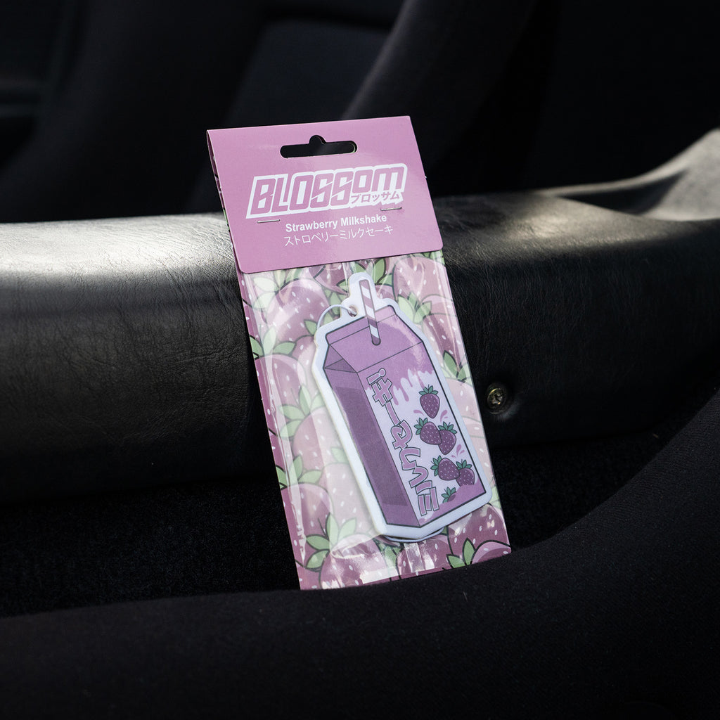A pink and purple 'The Milkshake Collection' Strawberry Milkshake air freshener by Blossom Automotive, shaped like a milk carton with Japanese text, sits on a car seat; its packaging features a strawberry pattern.