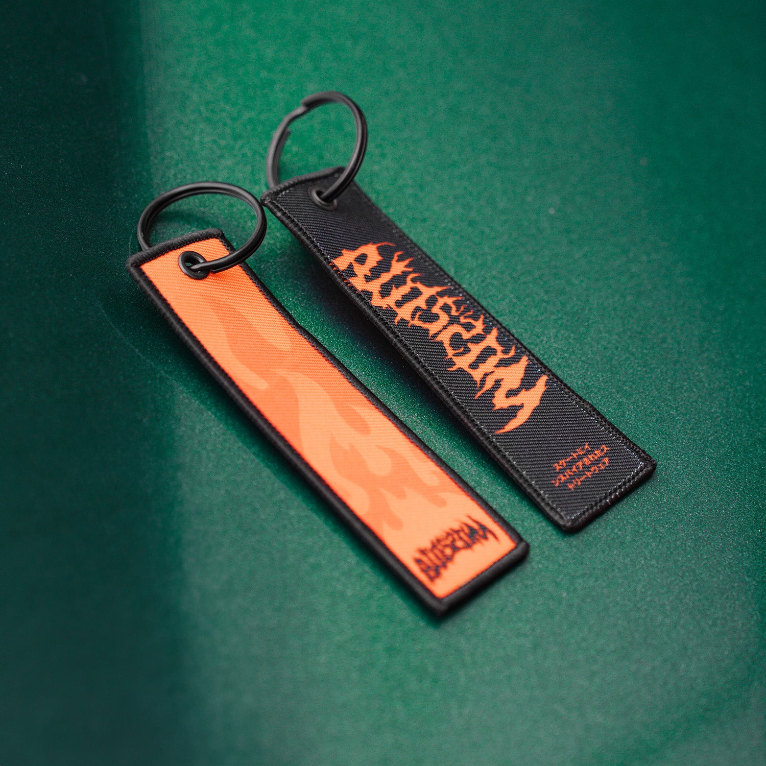 Two 'The Skate Collection' Flight Tag keychains by Blossom Automotive rest on a green surface: one is orange with a flame pattern, the other is black with bold orange "AXISTORM" text. Both feature black key rings.