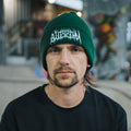 A man with a mustache and goatee, wearing Blossom Automotive's 'The Skate Collection’ Beanie in bottle green and a black shirt, looks directly at the camera inside an indoor skate park.