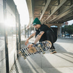 A skateboarder in Blossom Automotive’s 'The Skate Collection’ Beanie - Bottle Green and black clothes performs a mid-air trick at an urban skatepark beneath an overpass, sunlight streaming through a fence.
