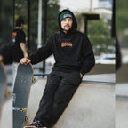 A man wears the Blossom Automotive ‘The Skate Collection’ Beanie in Bottle Green, sitting on a skatepark ramp with his skateboard. He gazes seriously at the camera; a blurred figure and greenery are visible in the background.