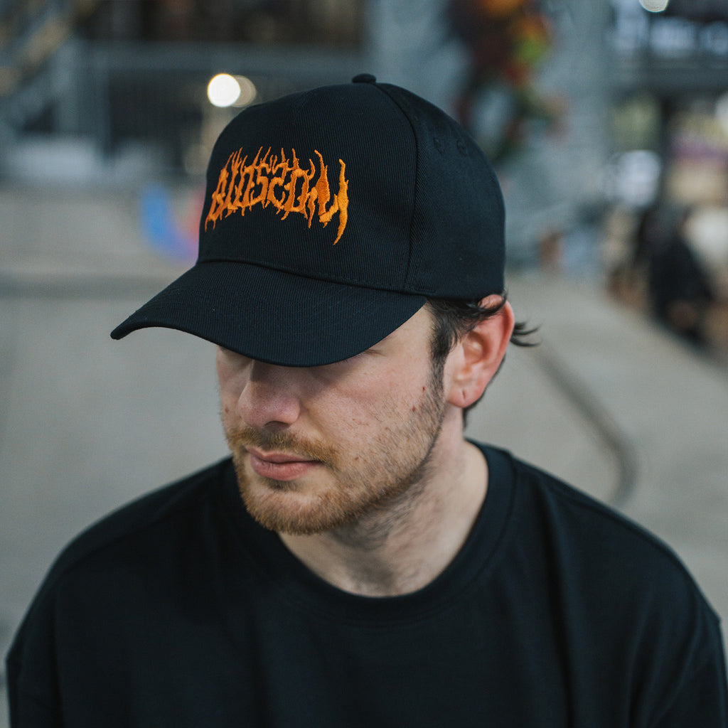 A man wears the Blossom Automotive 'The Skate Collection’ Cap in black and a matching shirt indoors, gazing slightly downward. The blurred background hints at an urban or skate park environment.