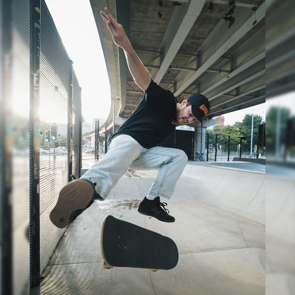 A skateboarder wearing the Blossom Automotive 'The Skate Collection’ Cap - Black does a trick mid-air at an outdoor skatepark under a bridge, sunlight streaming through a nearby fence as his board flips beneath him.