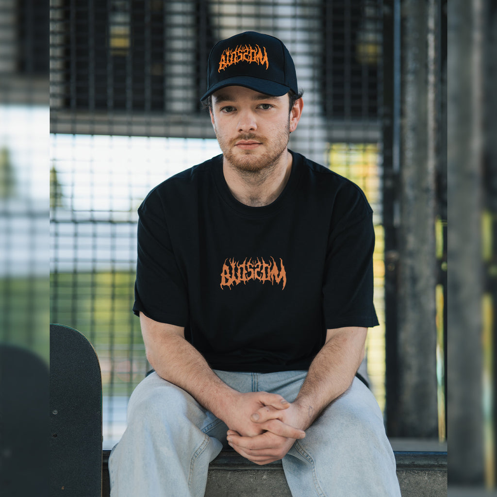Wearing Blossom Automotive’s ‘The Skate Collection’ Cap in black, a person sits on a bench with hands clasped, their skateboard nearby and a metal fence visible in the background.