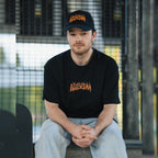 Wearing Blossom Automotive’s ‘The Skate Collection’ Cap in black, a person sits on a bench with hands clasped, their skateboard nearby and a metal fence visible in the background.