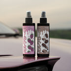 Two 200ml Blossom Automotive “The Milkshake Collection” car spray air fresheners sit on a car hood—one labeled Strawberry Milkshake with strawberries, the other Cookies & Cream. Both feature illustrated labels and the “It’s Qute” design.