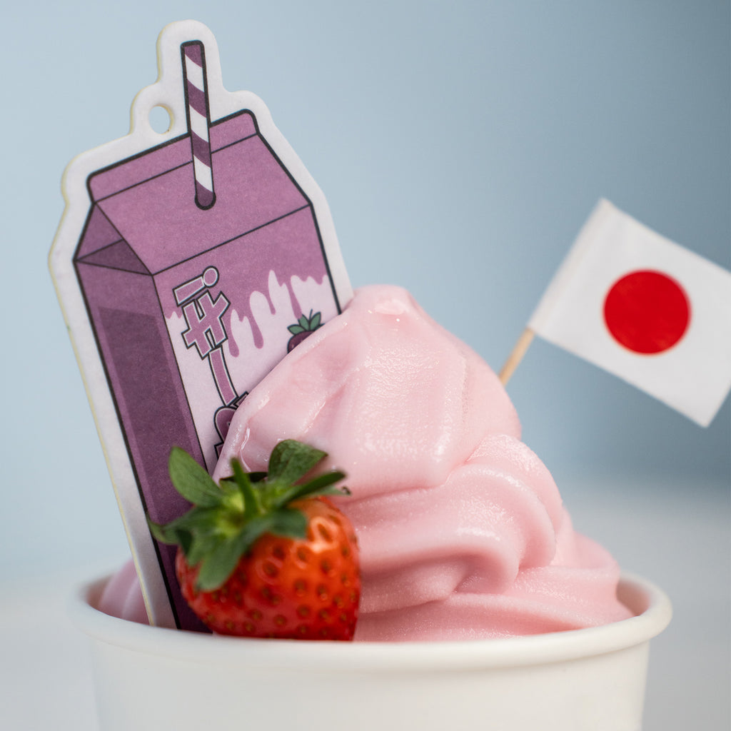 A 'The Milkshake Collection' Air Freshener - Strawberry Milkshake by Blossom Automotive, shaped like pink soft-serve with a strawberry, Japanese flag, and cute purple milk carton stick, set against a light blue background.