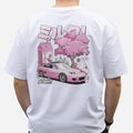 A person wearing the Blossom Automotive Milkshake Collection Oversized T-Shirt in white, featuring a pink cartoon graphic with a sports car, cherry blossom tree, milk carton, and Japanese text on the back.