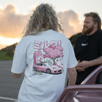 A person with curly blond hair wears Blossom Automotive's The Milkshake Collection Oversized T-Shirt in white, featuring a pink car, cherry blossom tree, and strawberry milk with Japanese text. Another person stands beside them near a parked car at sunset.