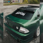 A green modified car parked on gravel features the Blossom Automotive Blossom Skate Logo XL Rear Window Die-Cut Sticker, a large white decal with graffiti-style text, with graffiti-covered walls and greenery in the background.