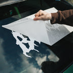 A person’s hand peels transfer paper to apply a white Blossom Skate Logo - XL Rear Window Die-Cut Sticker from Blossom Automotive onto a car’s rear window.