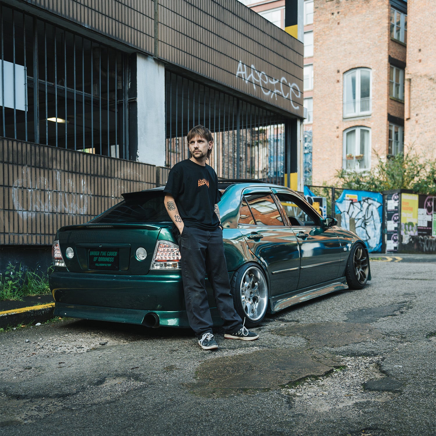 Model leaning on green IS200, wearing Blossom Skate Collection black tee, featuring Lexus IS200 graphic, oversized fit, inspired by skate and drift culture