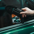 A hand uses a black squeegee labeled Blossom Automotive to apply The Skate Collection - Slap Sticker, featuring a racing car with flames and mountains, to a green car window.