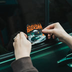 A person places The Skate Collection - Slap Sticker by Blossom Automotive, featuring graffiti-style text and an illustrated car scene, on the window of a green car.