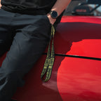 A person in black pants and a black shirt leans against a shiny red car, holding Blossom Automotive’s ‘The Mx5 Collection’ yellow and black lanyard, with their left hand in their pocket and wearing a black wristwatch.