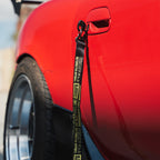 Close-up of a red car’s side with a wide rear tire and ‘The Mx5 Collection’ Lanyard by Blossom Automotive, featuring “GReddy x 9K” and “Automotive Apparel,” attached to the door handle.