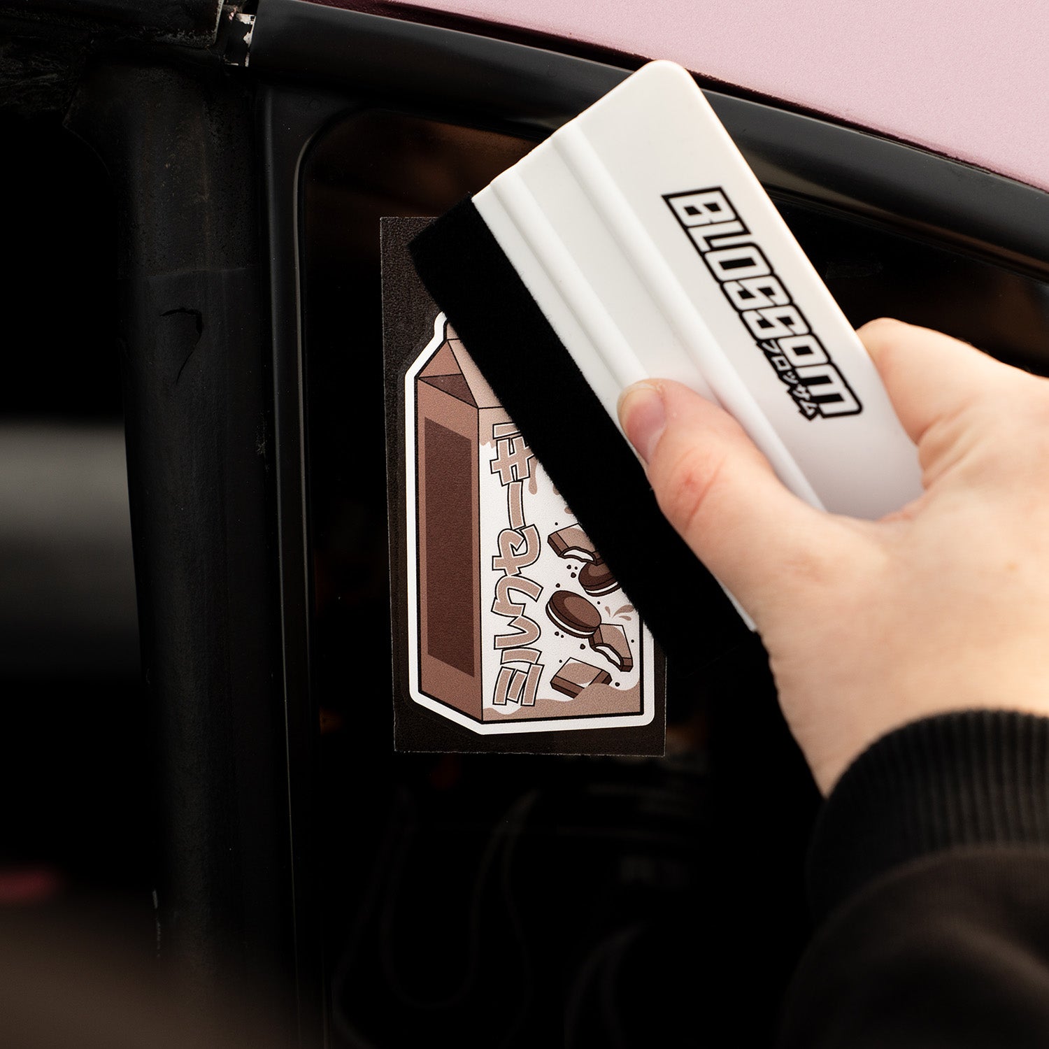 A hand uses the Blossom Automotive Blossom Felt Edge Application Squeegee - White to apply a chocolate milk carton “SLAP” sticker with cartoon splashes onto a car window.