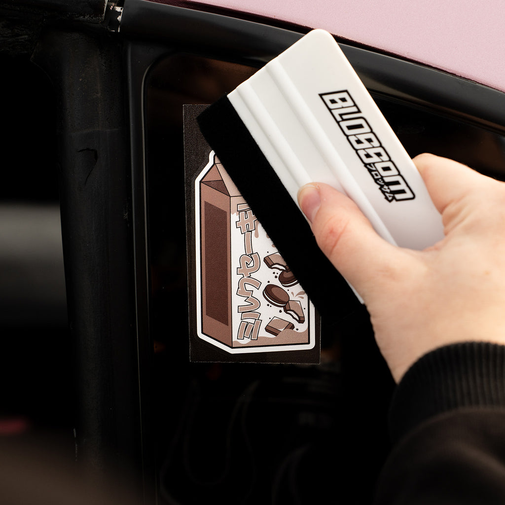 A hand applies the Blossom Automotive Cookies and Cream Milkshake - Slap Sticker to a car window using a white plastic squeegee.