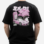A person wearing a black t-shirt with a graphic design featuring a pink car, strawberry milkshake and cherry blossoms on the back.