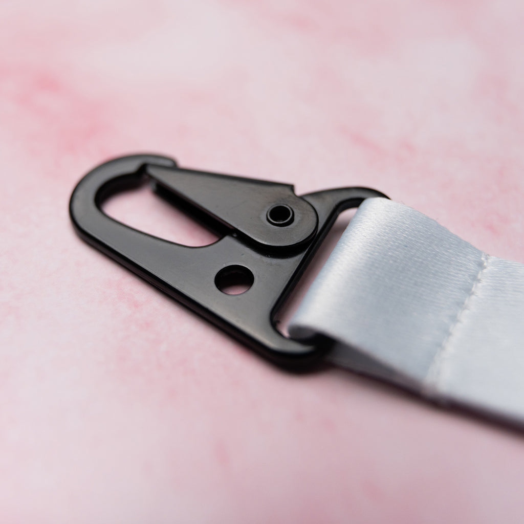 A close-up of ‘The Milkshake Collection’ Lanyard by Blossom Automotive features a black metal swivel hook clasp attached to a white strap, set against a soft pink background.