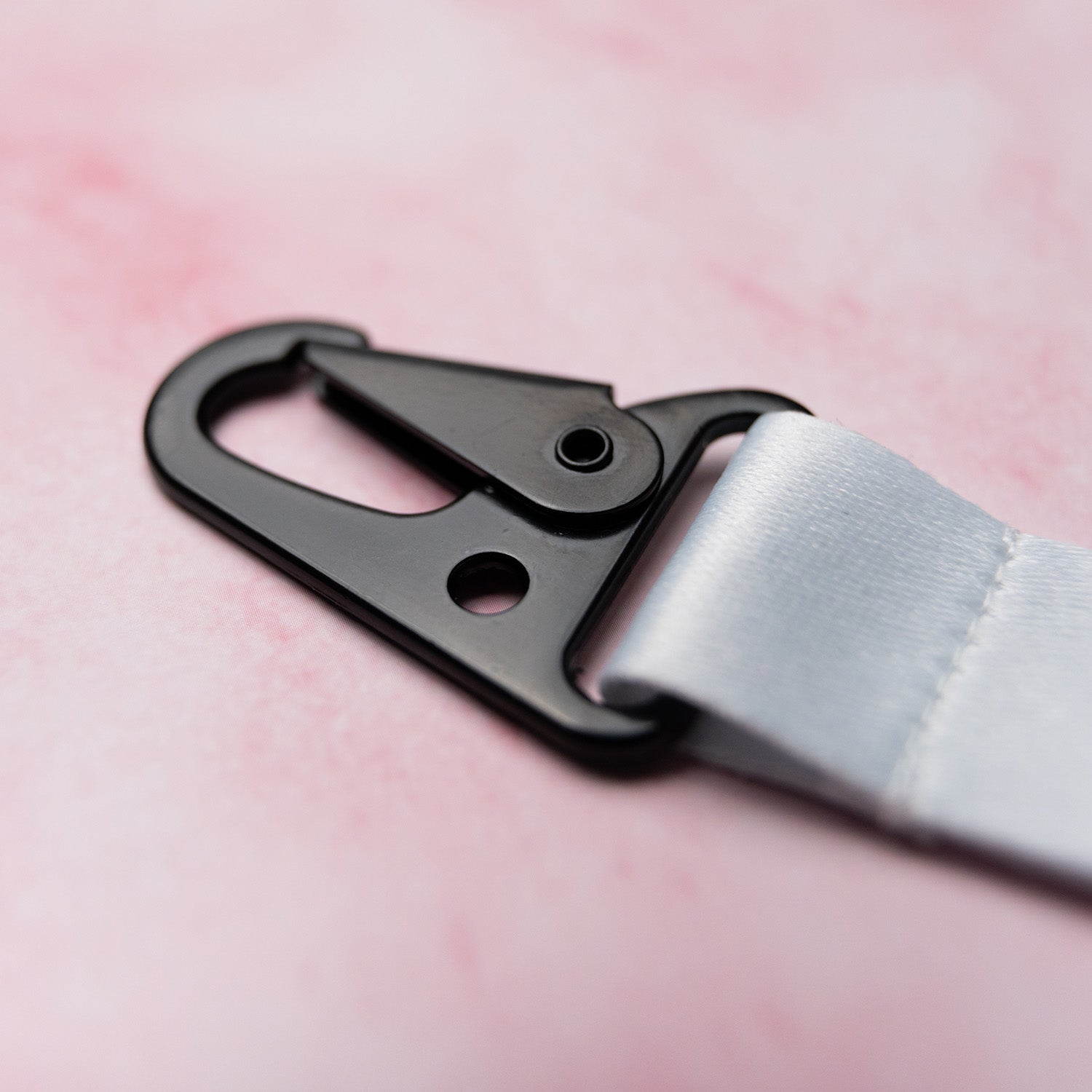 A close-up of ‘The Milkshake Collection’ Lanyard by Blossom Automotive features a black metal swivel hook clasp attached to a white strap, set against a soft pink background.