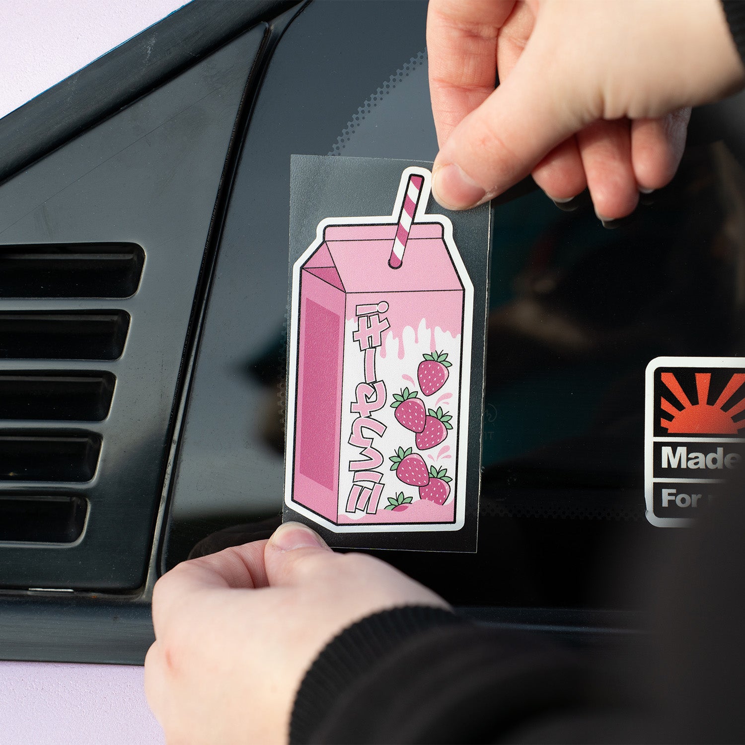 Someone sticks a Blossom Automotive Strawberry Milkshake - Slap Sticker featuring pink hues and Japanese text with strawberries onto a car window near the black vent.