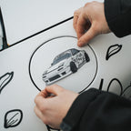 A person applies the Blossom Automotive Mx5 - Slap Sticker to the fuel door of a white car decorated with black line art decals.