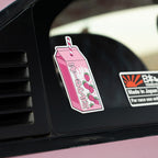 A Blossom Automotive Strawberry Milkshake - Slap Sticker, featuring a pink carton with Japanese text and strawberries, is displayed on a car window next to a Made in Japan sticker.