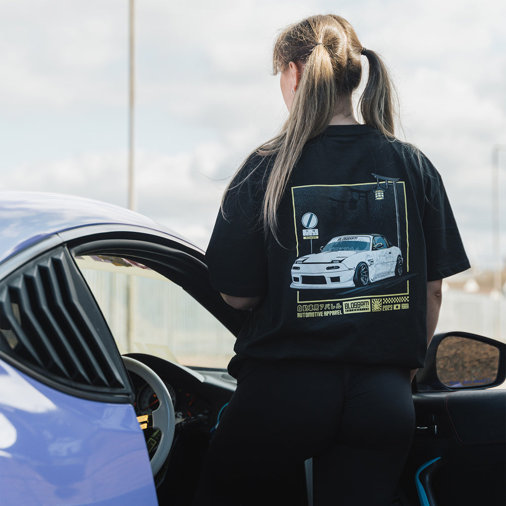 A person standing next to a car, wearing black crewneck t-shirts with a yellow graphic design on the back, featuring a white mx5.