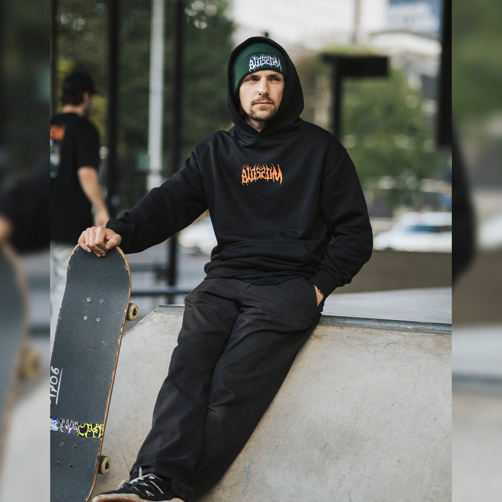 a person sitting in a skate park holing a skateboard, wearing a black skate collection hoodie and a green beanie