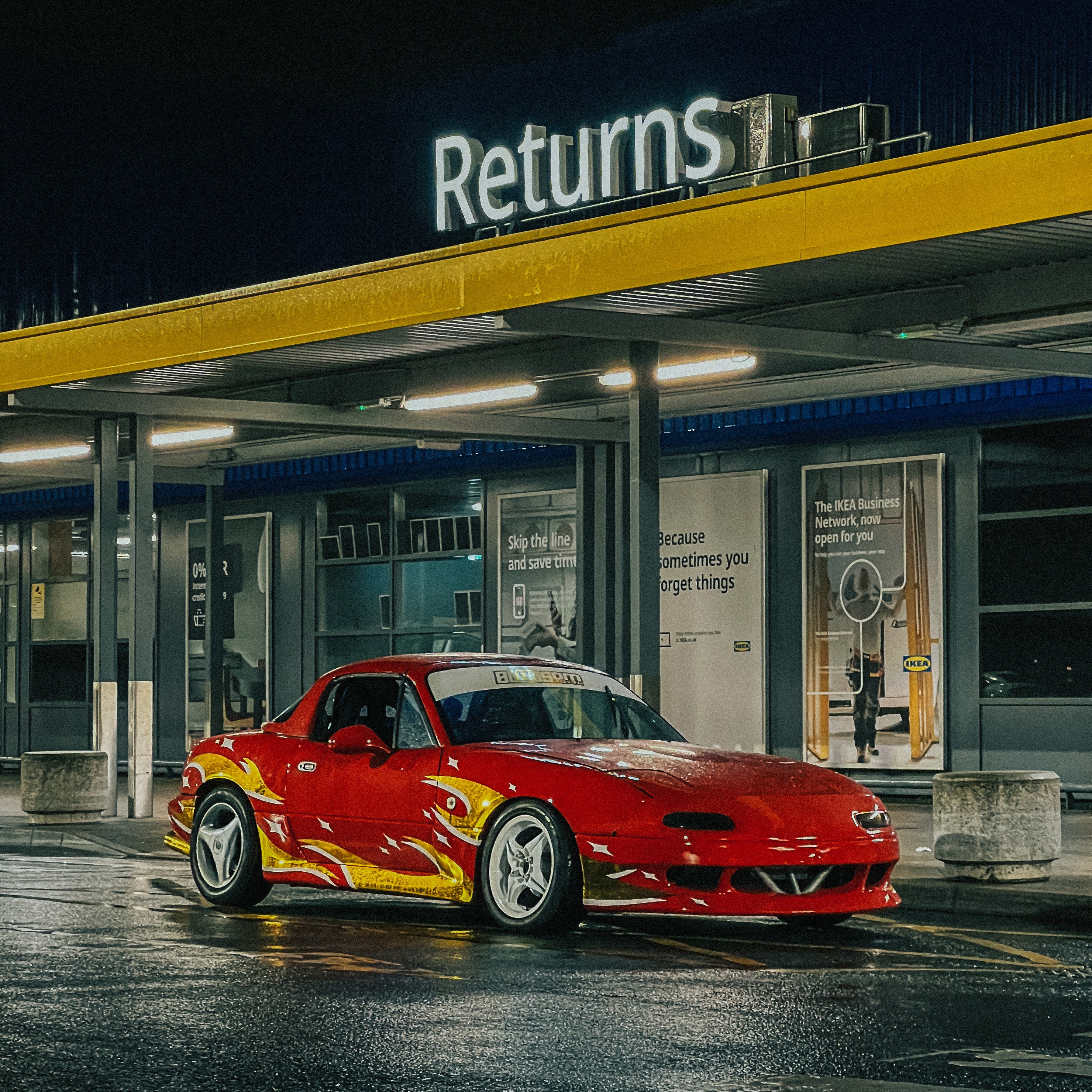A car with Blossom Automotive's DIY 'Zoku Style' Tribal Universal Customisable Hood Decals is parked at night in front of a store returns area, bright yellow lights and wet pavement reflecting the scene.