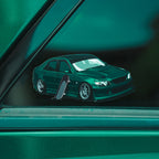 The IS200 - Slap Sticker by Blossom Automotive, featuring a green modified car with a skateboard, is displayed on the window of a green vehicle. Both the sticker and car share similar green shades.