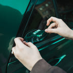 Someone carefully applies the Blossom Automotive IS200 - Slap Sticker to the small triangular window of a green car, aligning it with both hands. The car's window reflects a nearby building.